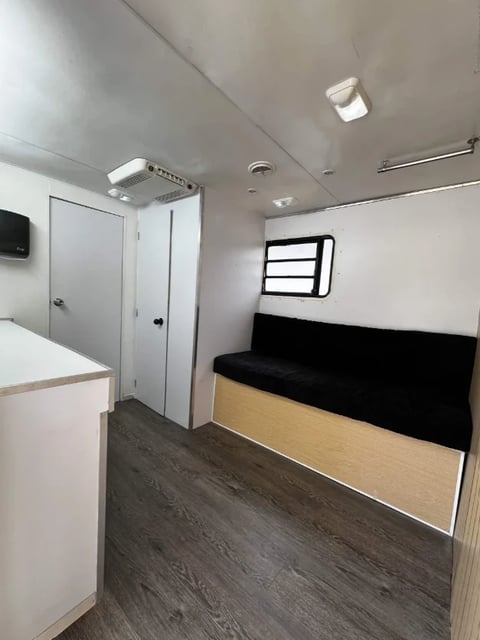 Interior of a small trailer or RV with white walls, a black cushioned bed on the right, wooden cabinetry, and a bathroom door on the left
