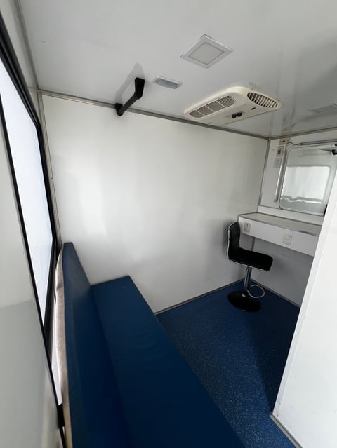Interior of a white and blue enclosed mobile office or utility trailer with ceiling fan, air conditioning unit, and desk workspace