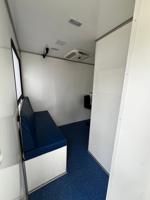 Small interior room with white walls, blue carpeted floor, blue bench seating, and ceiling vents visible from doorway entrance
