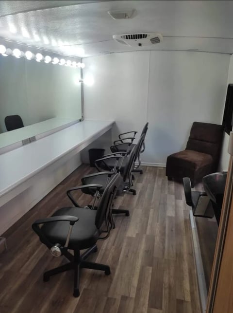 Interior of a mobile trailer with makeup vanity station featuring mirrors, LED lights, black chairs, and brown armchair
