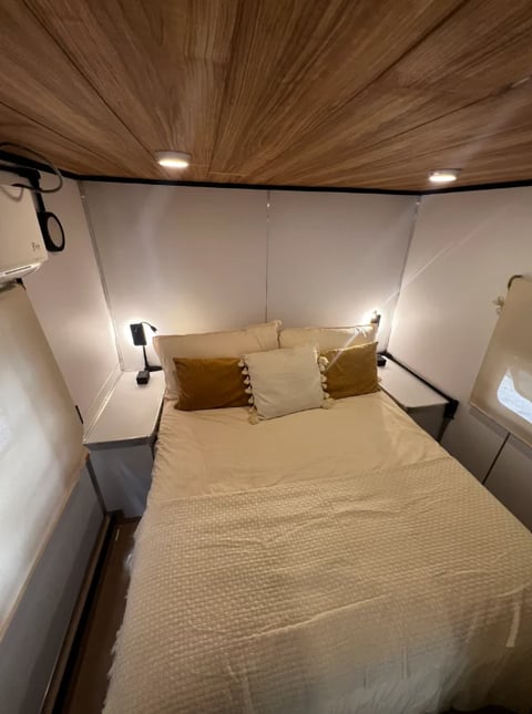 Modern RV bedroom with queen bed, wood ceiling, ambient lighting, and tan pillows and bedding