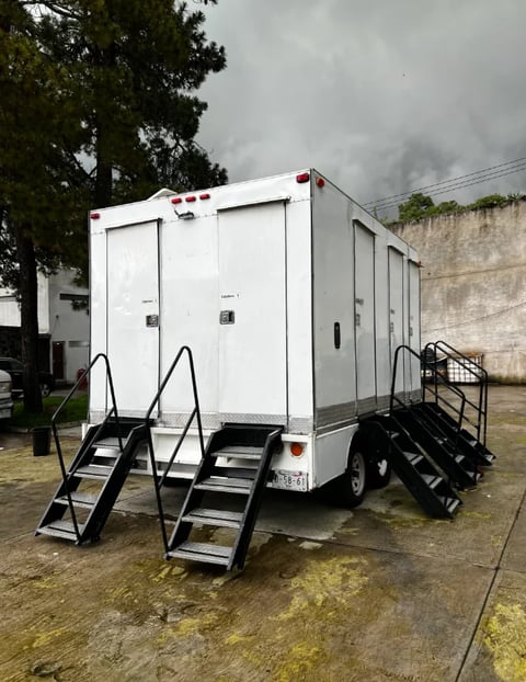 White portable restroom trailer with multiple stalls and black metal stairs on either side, parked on concrete with trees and overcast sky in background