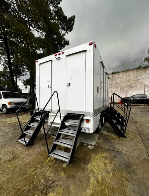 White portable restroom trailer with black metal stairs and handrails parked on wet ground near trees