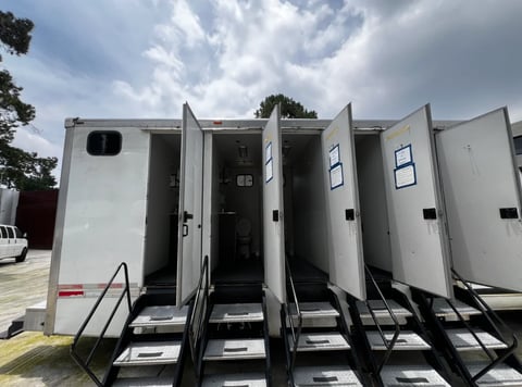 White mobile restroom trailer with multiple open doors and entry stairs under cloudy sky