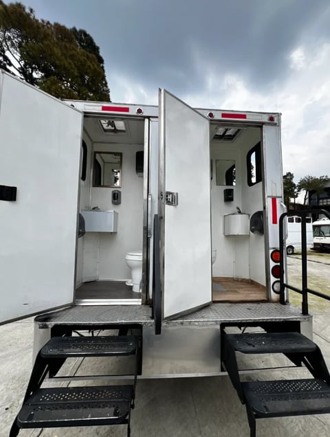 Two white portable toilet units with open doors, black step platforms, and red trim accents parked outdoors