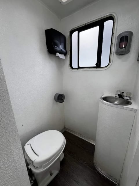 Compact RV bathroom with white walls, toilet, sink, window, and wall-mounted storage compartments