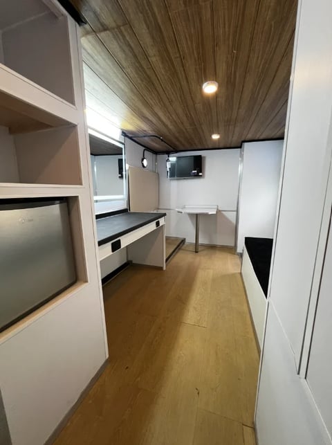 Interior of a modern RV or camper showing kitchen area with white cabinets, wood ceiling, and compact appliances along wooden flooring.