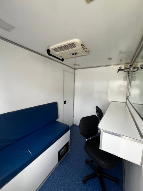 Interior of a small office or mobile workspace with blue bench seating, black chair, white desk, ceiling air conditioning unit, and blue flooring