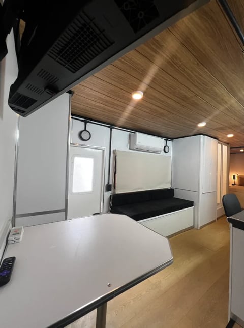 Modern RV interior with white cabinets, wood ceiling, dining table, and sleeping quarters with black cushioned bed