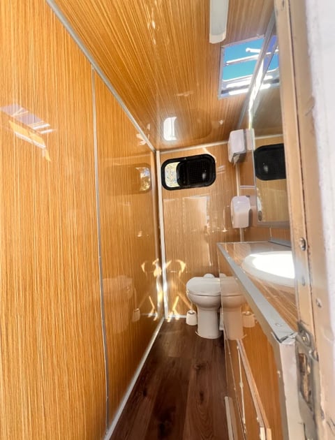 Interior of a narrow RV bathroom with wood paneling, toilet, sink, and glass shower enclosure with blue exterior visible through window