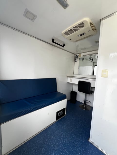Interior of a compact RV or mobile office with blue seating, white desk area, and ceiling-mounted air conditioning unit