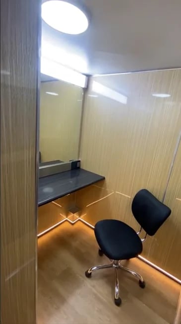 Small office booth with beige walls, bright overhead lighting, black desk, and rolling chair with warm floor lighting