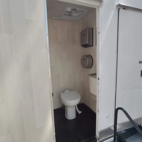 Interior view of a portable toilet stall with white pedestal toilet, dispensers, and recessed ceiling mounted in a small compartment
