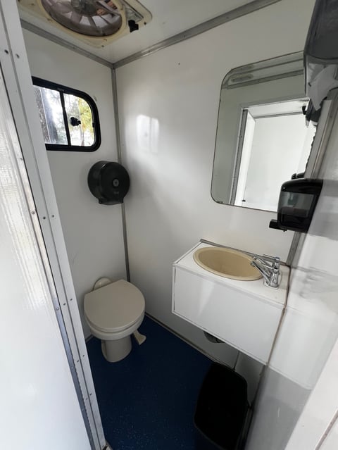 Interior of a compact RV bathroom with toilet, sink with mirror, small window, and overhead ventilation fan