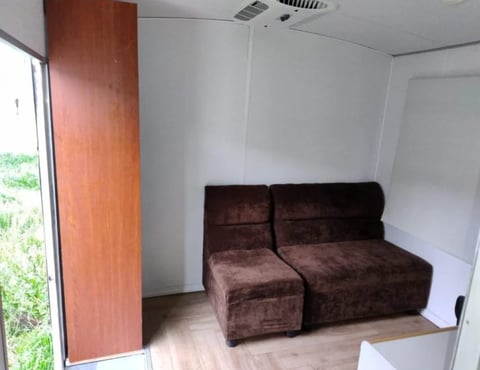 Interior of compact mobile living space with brown upholstered seating, wooden door on left, white walls, and light wood flooring.