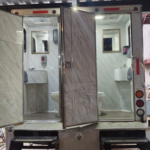 Two portable bathroom units with gray tile interiors, sinks, and toilets, displayed side by side with open doors