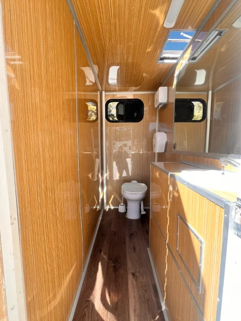 Interior of an RV or travel trailer with wood grain paneling, showing toilet, sink, narrow hallway, and skylights overhead