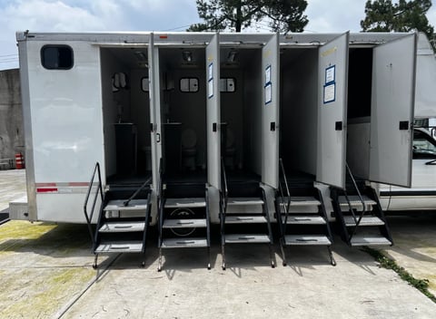 White portable restroom trailer with five individual stall doors and metal stairs on outdoor ground