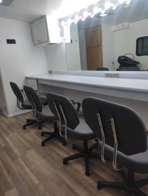 Professional vanity station with three black office chairs, large mirror with bright bulb lights, and wood flooring in a modern salon or makeup room