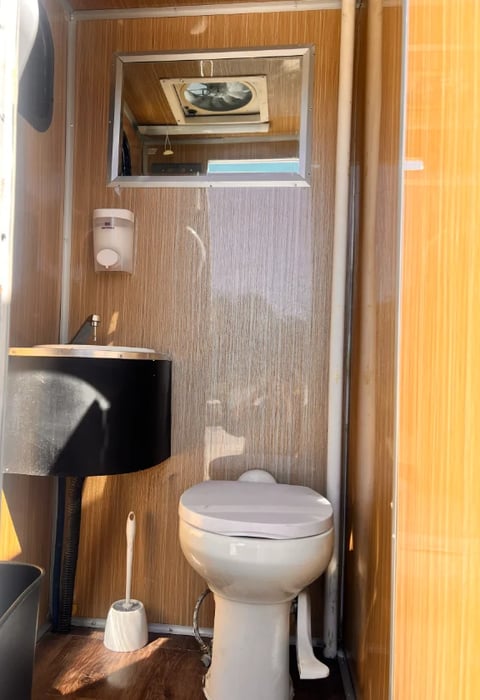 Compact RV bathroom with toilet, small black sink, toilet brush, and storage shelf with books and supplies