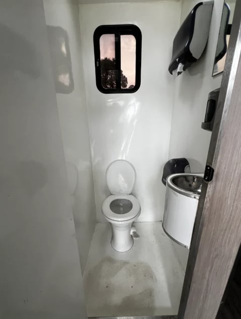 Interior of an RV bathroom with white walls, a small window, white toilet, black fixtures on the wall, and a trash can on the right side