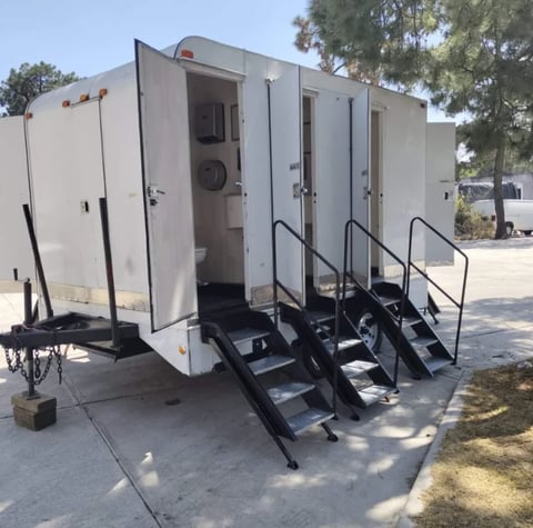 White mobile restroom trailer with four black metal stairs and handrails parked outdoors in daylight