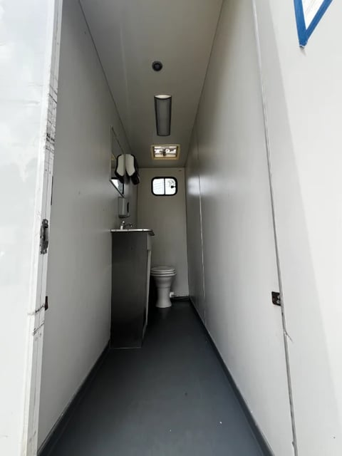 Interior view of a narrow portable restroom or trailer with white walls, black floor, sink, toilet, and overhead lighting