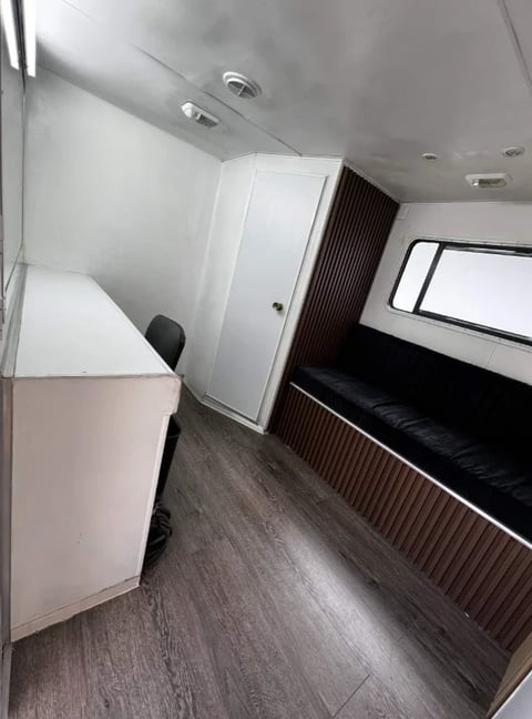Interior of an RV or trailer showing white cabinetry, black bench seating, wood-grain flooring, and ceiling fixtures