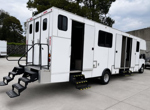White commercial vehicle with black-framed windows and doors, equipped with exterior stairs and wheelchair ramp on a concrete lot