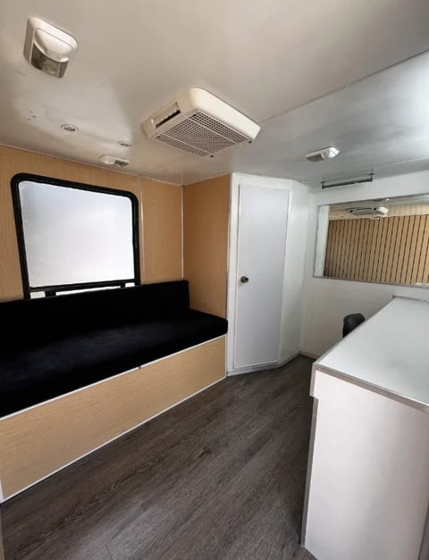 Interior of a small RV or trailer with wooden cabinets, a bed with black mattress, white appliances, and climate control units on ceiling