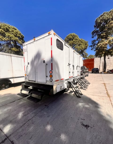 White enclosed cargo trailer with red stripe parked in lot, bikes mounted on rear ramp, trees and clear sky visible