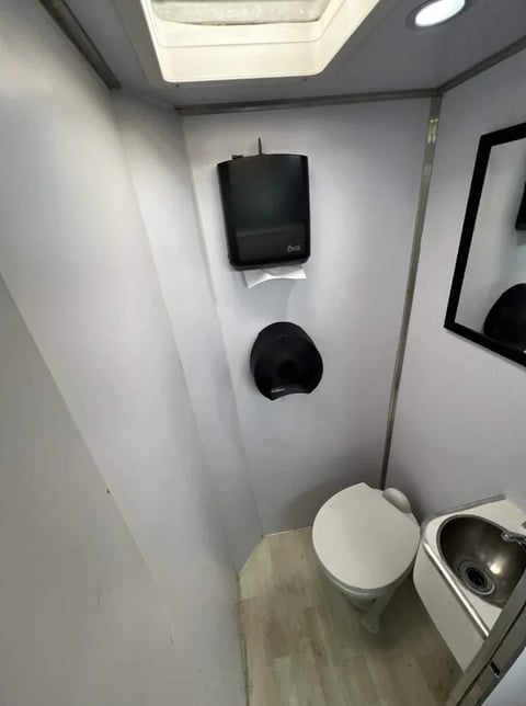 Interior of a portable toilet with white walls, white toilet, paper towel dispenser, and skylight ceiling